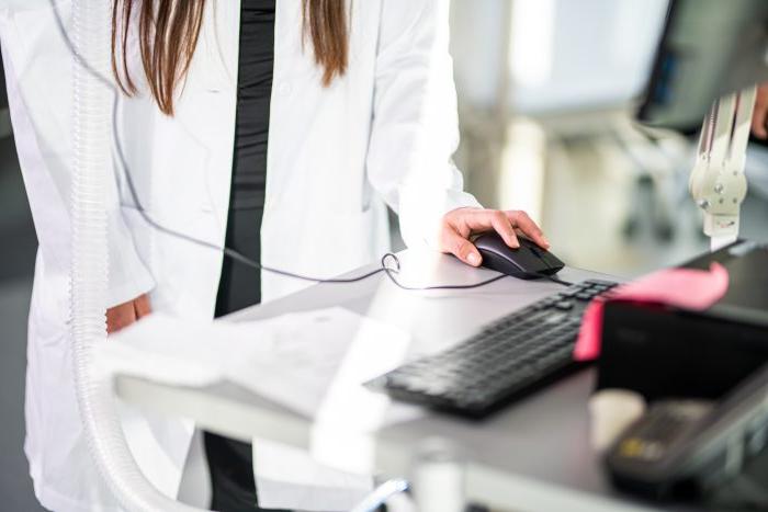 A person in a lab coat operates a computer mouse while standing at a workstation, with a keyboard and monitor visible on the desk.