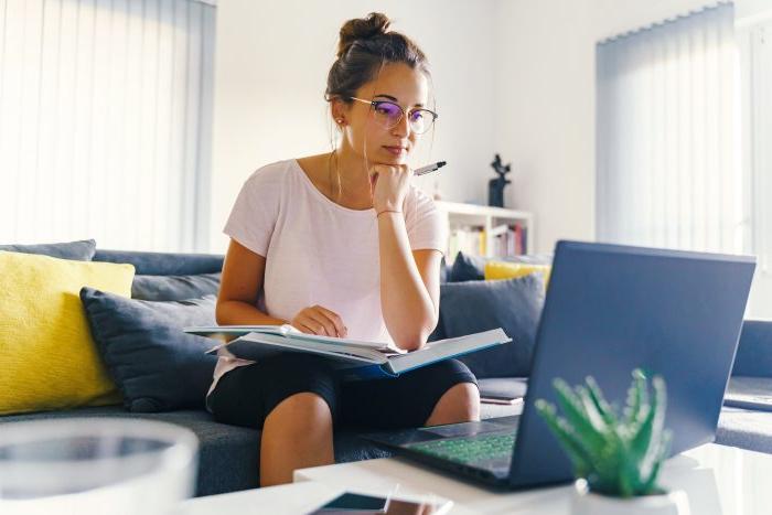 An adult learner sits on a couch in front of a laptop. Her chin is propped on her fist and she has a textbook and paper on her lap.