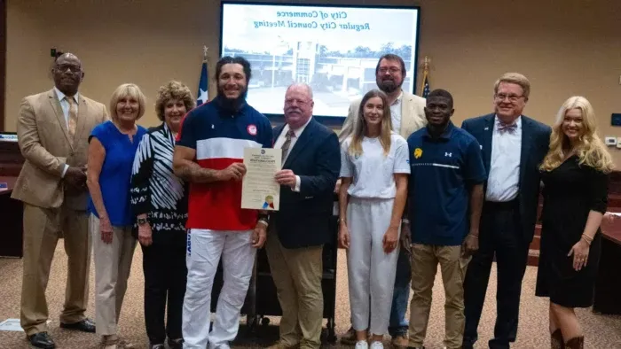 Ten individuals face the camera and smile, with a screen in the background that reads "City of Commerce Regular City Council Meeting."